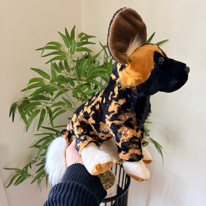 A plush toy dog with a black and tan spotted pattern, large upright ears, a white tail, and a black face, being held up by a person's hand in front of a potted green plant against a light wall.