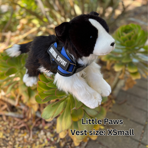 Black and white stuffed dog toy with a blue vest labeled 'Little Paws' on green plants.
