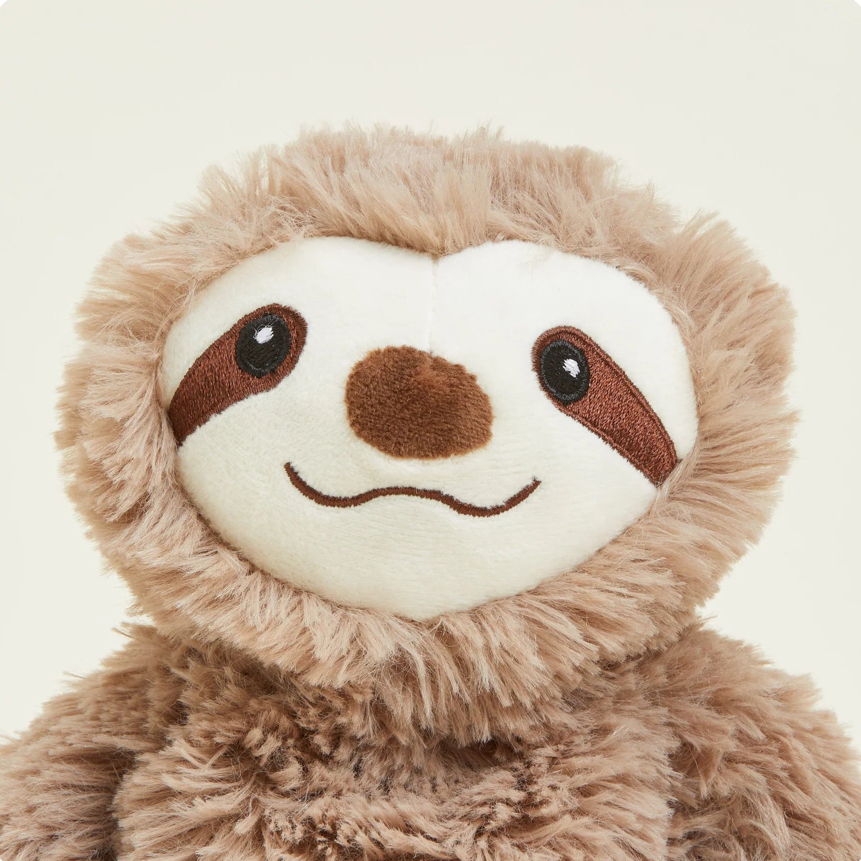 A close-up of a plush sloth toy with fluffy light brown fur and a cream-colored face, featuring embroidered brown eyes, nose, and mouth details, set against a solid off-white background.