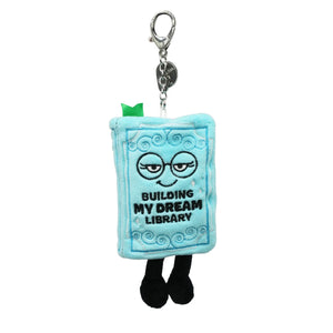 A plush book-shaped keychain charm with a silver metal clasp. The light blue book has embroidered black text reading 'BUILDING MY DREAM LIBRARY' and a simple face, with small black legs and a tiny green accent on top.