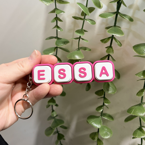 Hand holding a keychain with the word 'ESSA' in pink and white letters against a green leafy background.