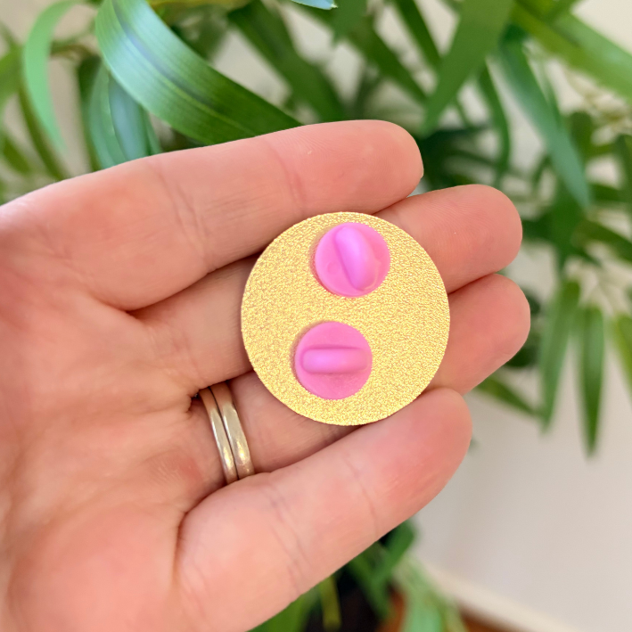 A hand holding a round gold glitter pin featuring two pink male urethra symbols, with a blurred green plant in the background.