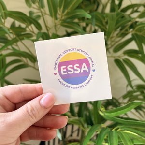 A hand holds a square white sticker featuring the ESSA logo, with a background of green houseplants.
