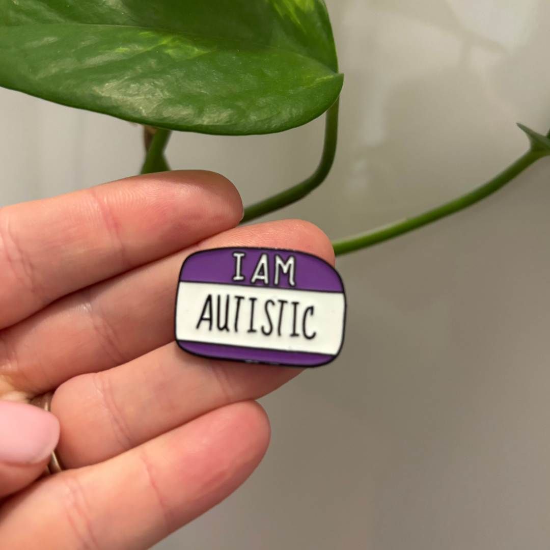 Hand holding a 'I AM AUTISTIC' badge with a green leaf background