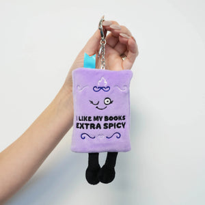 A hand holding a small plush book keychain in light purple. The plush has embroidered facial features, the text 'I LIKE MY BOOKS EXTRA SPICY' in black, blue decorative swirls, and black legs, attached to a silver metal key ring.