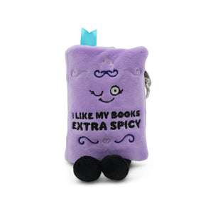A purple plush book-shaped keychain or pouch with embroidered details, including a smiling face, the text 'I LIKE MY BOOKS EXTRA SPICY', black feet, and a small light blue ribbon at the top, shown on a white background.