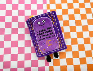 A purple plush book toy with embroidered cartoon eyes and the text 'I LIKE MY FANTASIES TO HELP ME ESCAPE REALITY' on a pink and white checkered background.