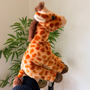 A plush giraffe toy being held up by a hand in front of a white wall and green potted plant. The toy has an orange and cream spotted pattern with brown accents on its mane, ossicones, and feet.