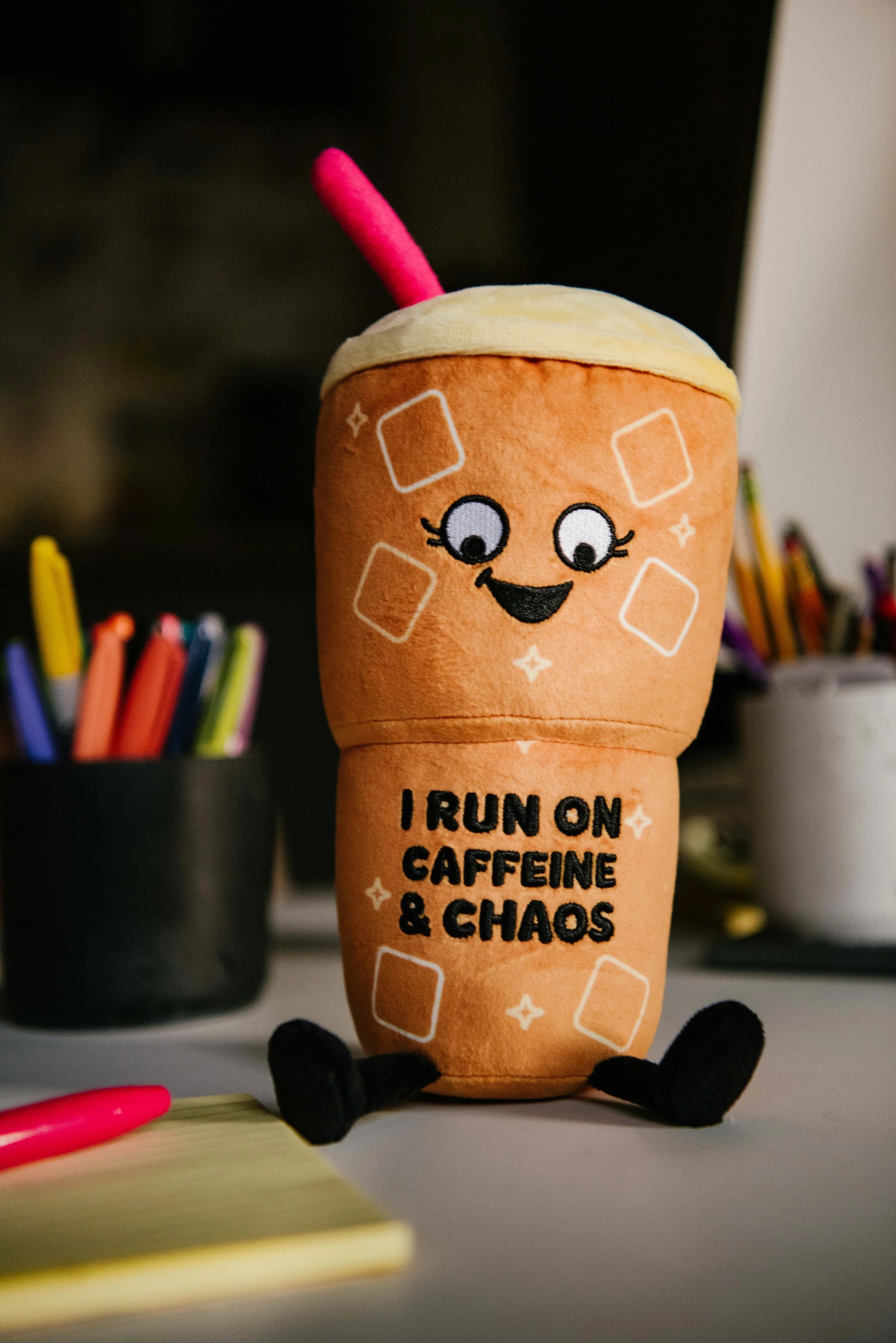 A plush toy shaped like a coffee cup with a smiling face, black legs, and a pink straw, sitting on a desk with school supplies in the background. The text on the cup reads 'I RUN ON CAFFEINE & CHAOS'.