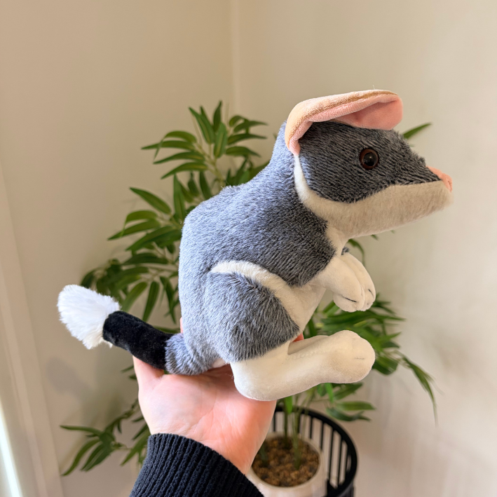 A plush toy mouse being held in a person's hand, with a potted plant and cream-colored wall in the background.