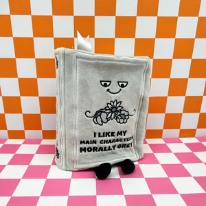 A plush gray book character toy with embroidered facial features, a flower motif, and the text 'I LIKE MY MAIN CHARACTER MORALLY GREW!' on the front, standing on a pink-and-white checkered surface against a matching orange-and-white checkered backdrop.