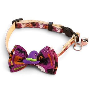 A pet collar with a decorative bow tie featuring a purple witch hat and a small green accent, attached to a patterned strap with a bell. Ghosts and pumpkins and black cats on collar pattern.
