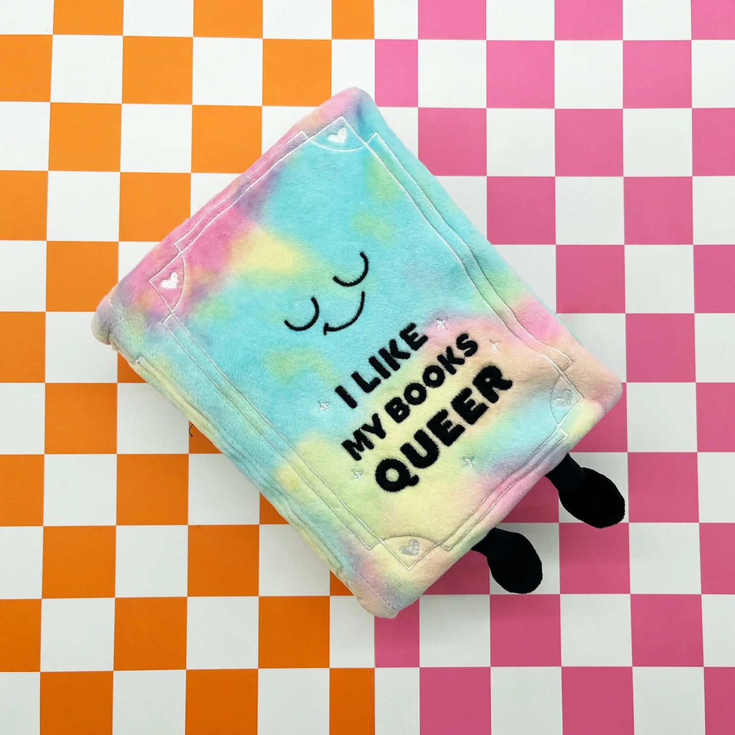 A pastel tie-dye plush book character with black legs and the text 'I LIKE MY BOOKS QUEER' printed on the front, placed on a pink and orange checkered background.