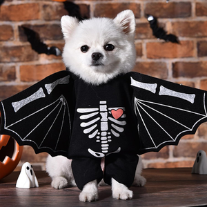 A small white dog wearing a black Halloween bat costume with wings and a white skeleton print with a red heart on the chest, standing on a wooden surface against a brick wall background.
