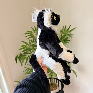 A plush black and white lemur toy being held up by a person's hand in front of a potted plant against a beige wall.