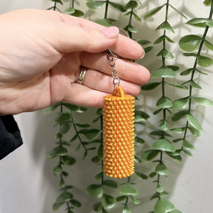 A hand holding a small orange textured cylindrical keychain or bag charm with a silver key ring, set against a background of hanging green ivy leaves.