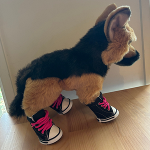 Stuffed dog toy with black and white shoes and pink laces on a wooden floor.
