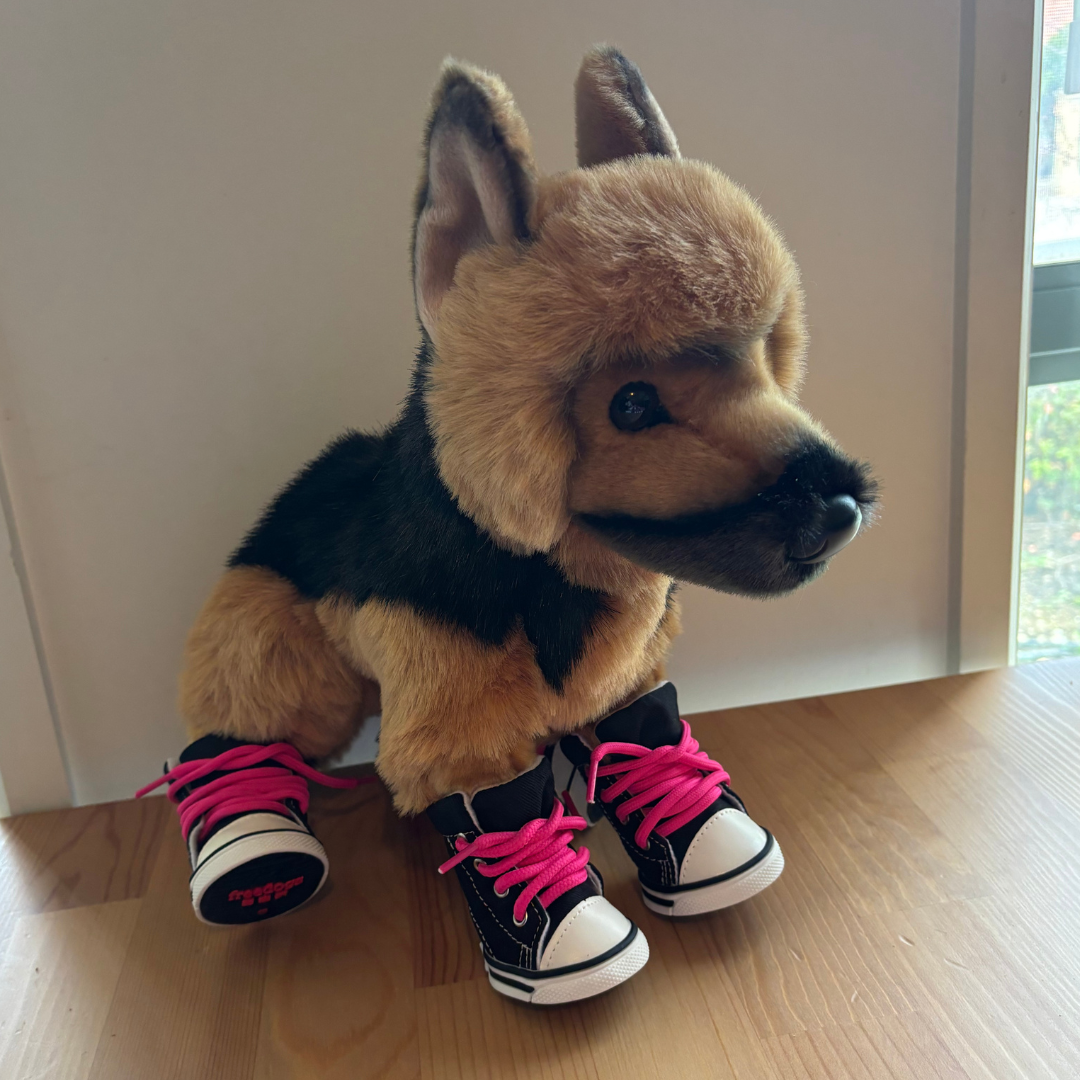 Plush dog toy wearing black shoes with pink laces on a wooden floor.