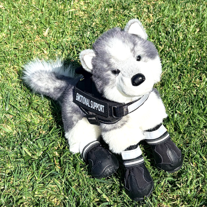 A plush stuffed dog toy dressed as a husky sits on green grass, wearing a black harness with the words 'EMOTIONAL SUPPORT' and black booties with white stripes.