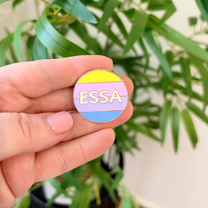 A hand holding a small round enamel pin with the word 'ESSA' in gold lettering across pastel yellow, pink, and blue horizontal stripes, with blurred green leaves in the background.