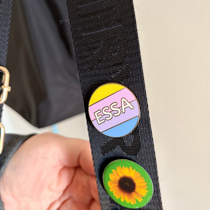 A close-up of a black woven strap with two oval pins attached. One pin is divided into yellow, pink, and blue stripes with the name 'ESSA' in white text.