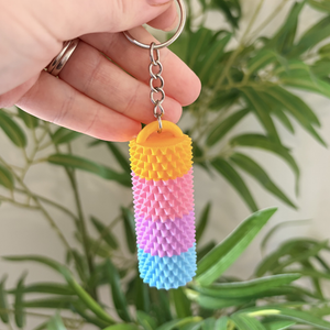 A hand holding a small textured keychain with a cylindrical, spiky design in yellow, pink, purple, and blue, with a silver key ring, against a blurred background of green houseplants.