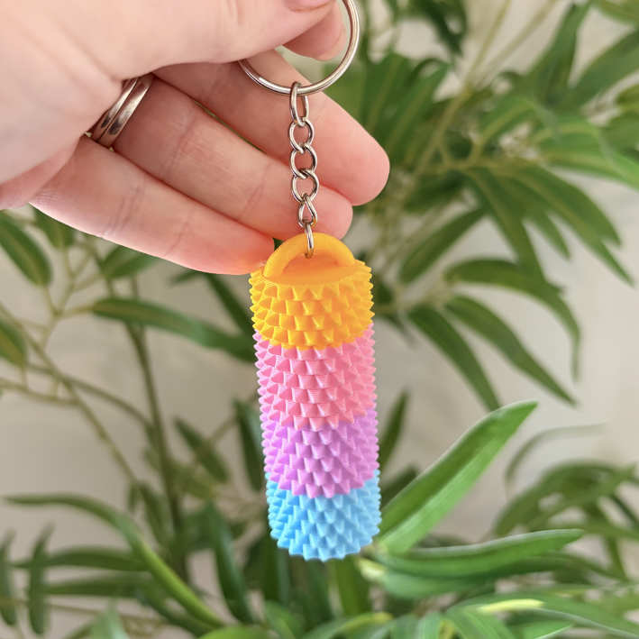 A hand holding a small textured keychain with a cylindrical, spiky design in yellow, pink, purple, and blue, with a silver key ring, against a blurred background of green houseplants.