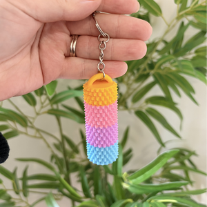 A hand holds a small cylindrical textured keychain with a yellow, pink, purple, and blue striped design, featuring a silver metal key ring and clasp, set against a blurred background of green houseplants.