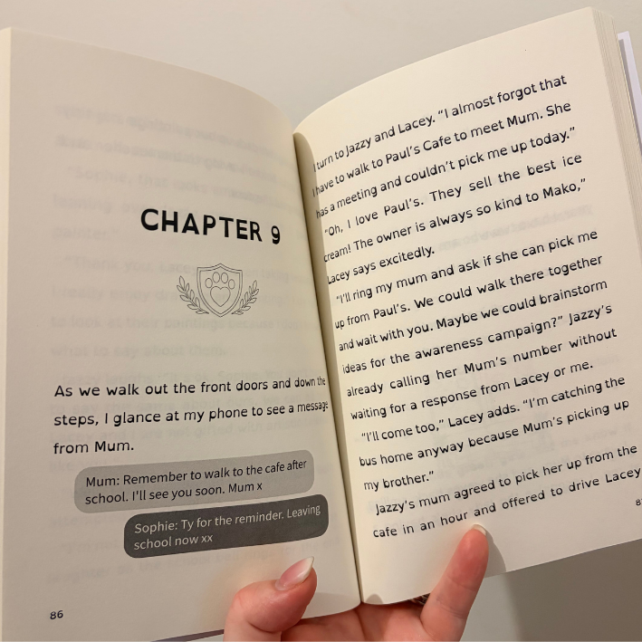 An open book showing printed text pages, with a hand holding the bottom edge. The left page has a message bubble and chapter heading 'CHAPTER 9', and the right page contains a narrative text passage.