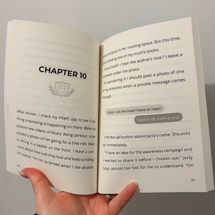 An open book held in a hand, showing two printed pages with text from a story or novel, including a chapter heading and dialogue bubbles.