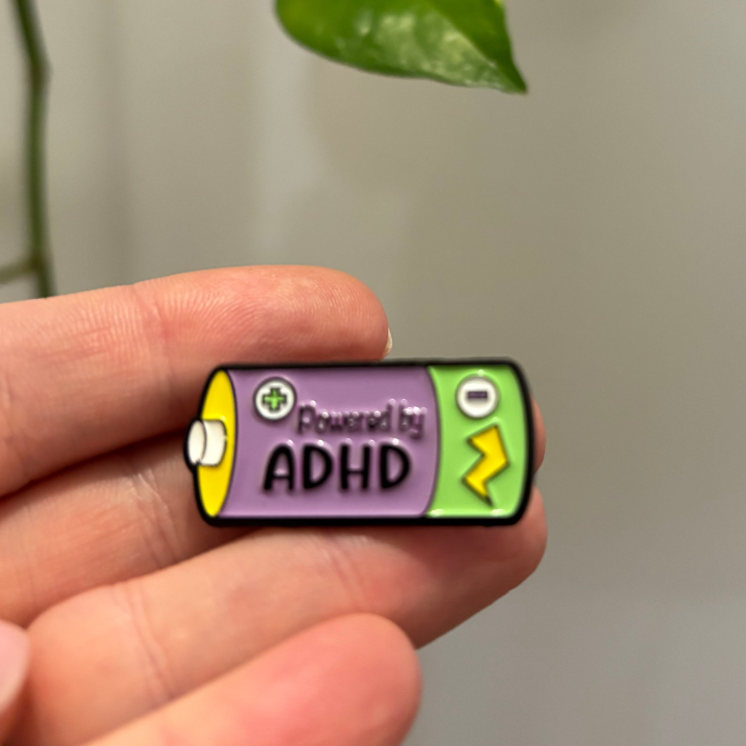 Pin with 'Powered by ADHD' design held between fingers against a neutral background