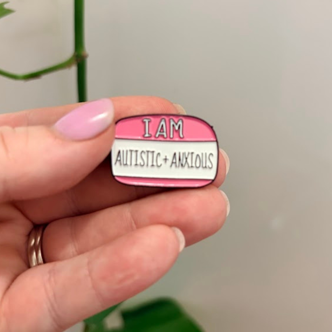 Pin with 'I AM AUTISTIC+ANXIOUS' text held by a hand against a neutral background