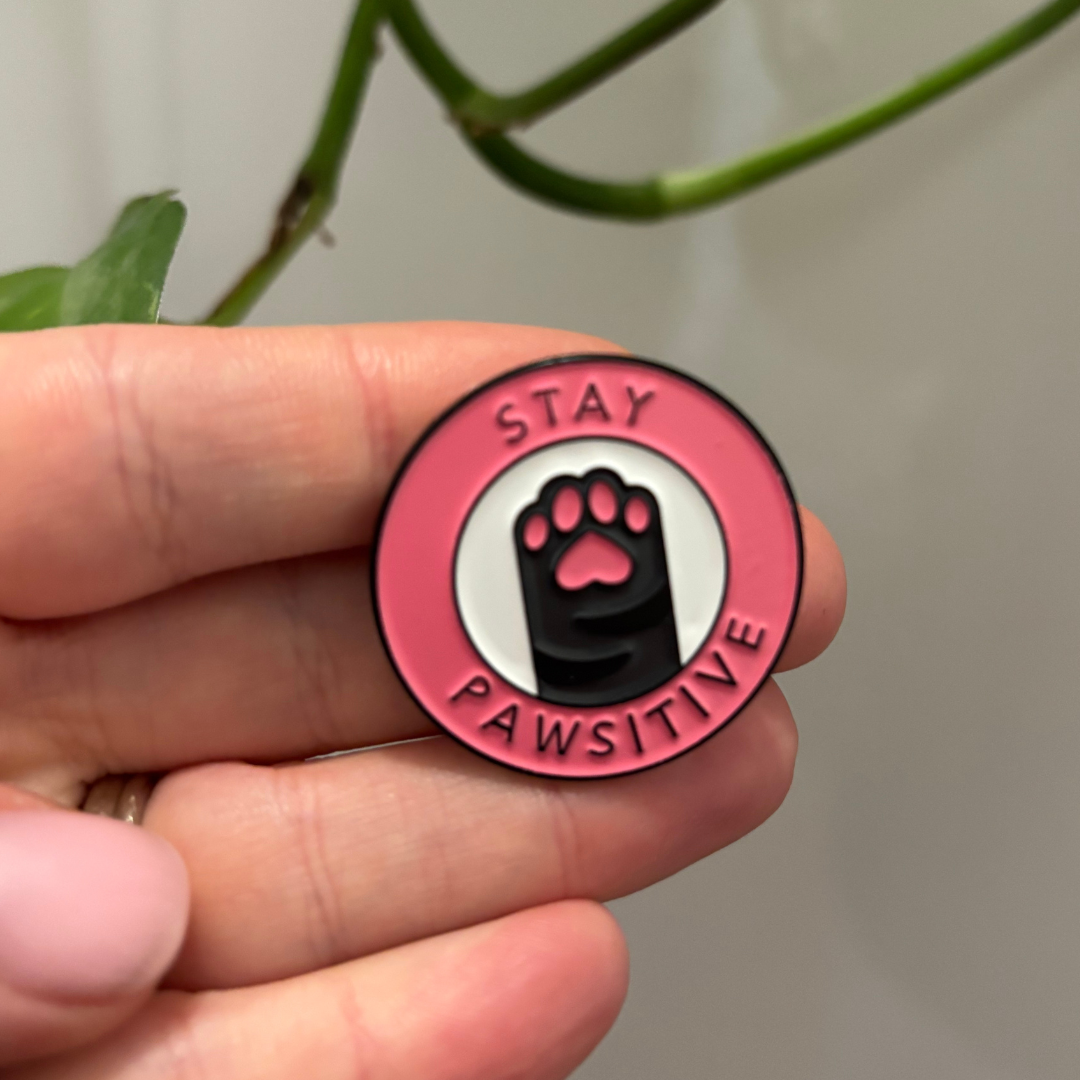 Pink enamel pin with paw print and 'Stay Pawsitive' text held in a hand.