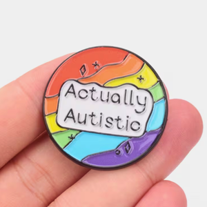 Colorful badge with 'Actually Autistic' text held in a hand against a white background