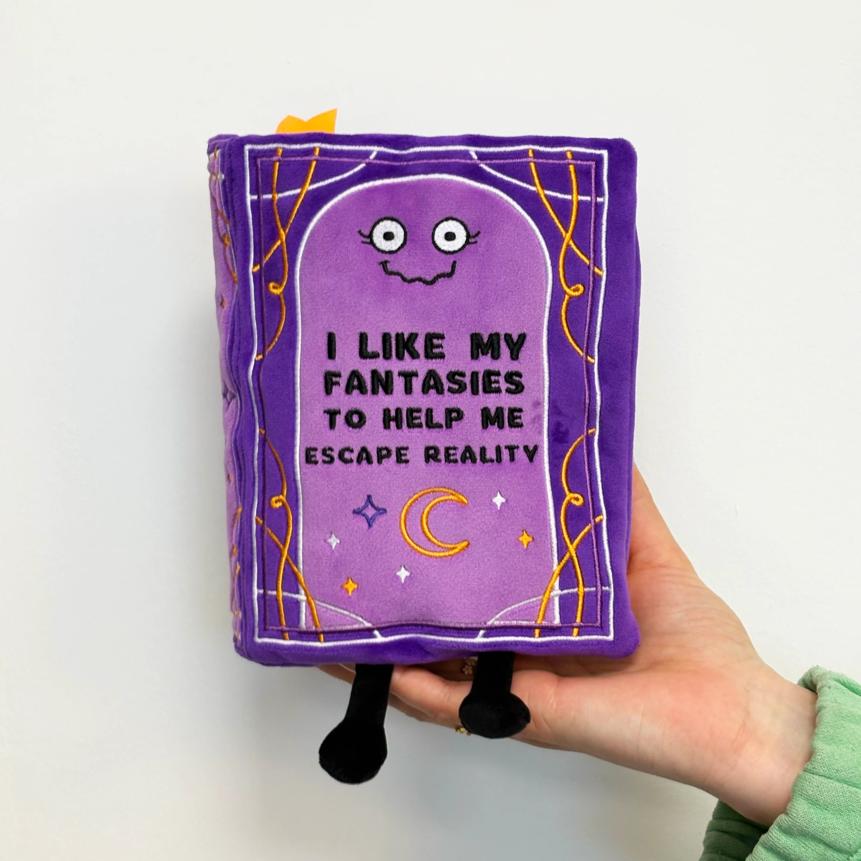 A hand holding a purple plush book toy with embroidered text that reads 'I LIKE MY FANTASIES TO HELP ME ESCAPE REALITY' and whimsical cartoon details, against a plain white background.
