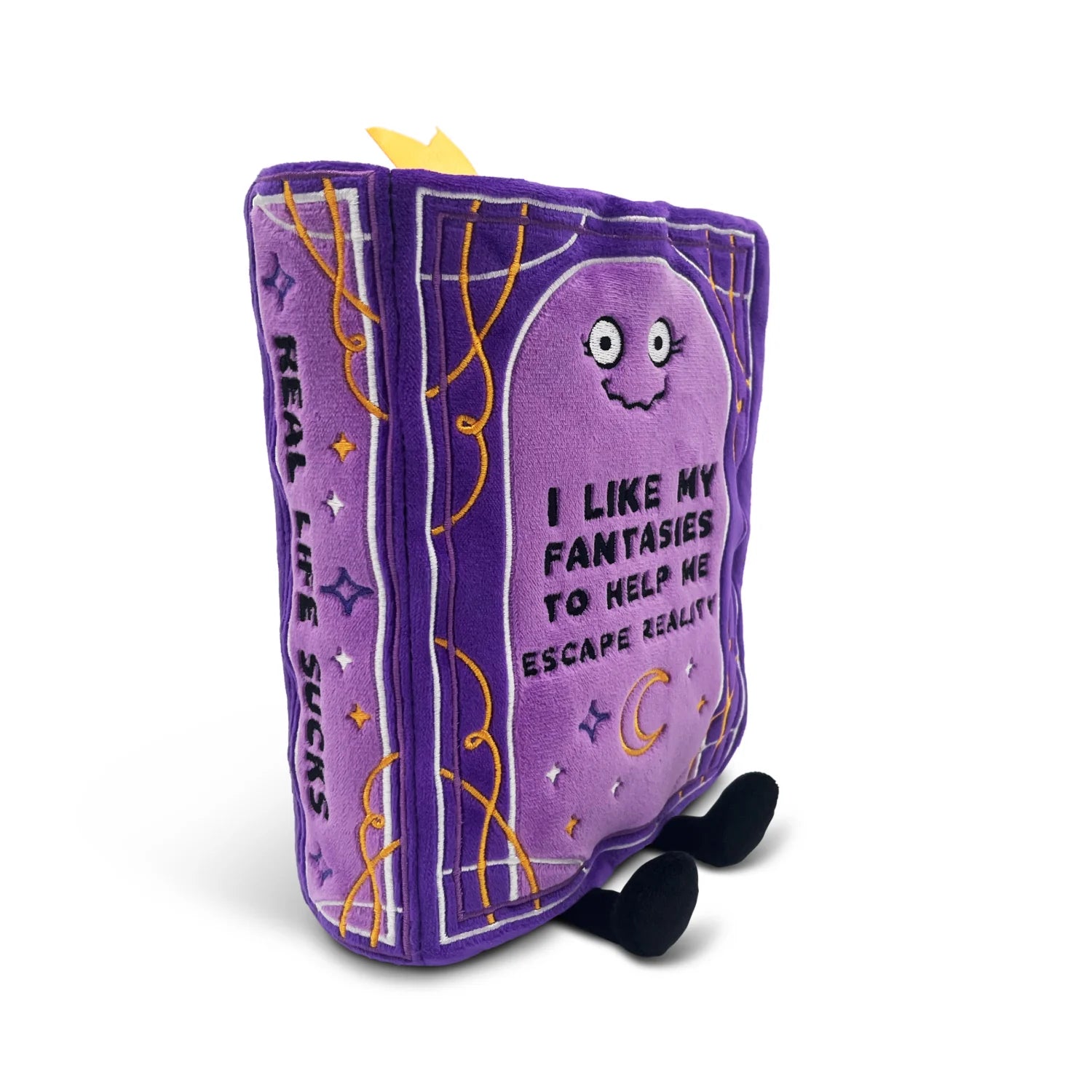 A purple plush book character toy with embroidered details, featuring the text 'I LIKE MY FANTASIES TO HELP ME ESCAPE REALITY' on the front cover, 'REAL LIFE SUCKS' on the spine, and small black feet and a yellow crown accent.