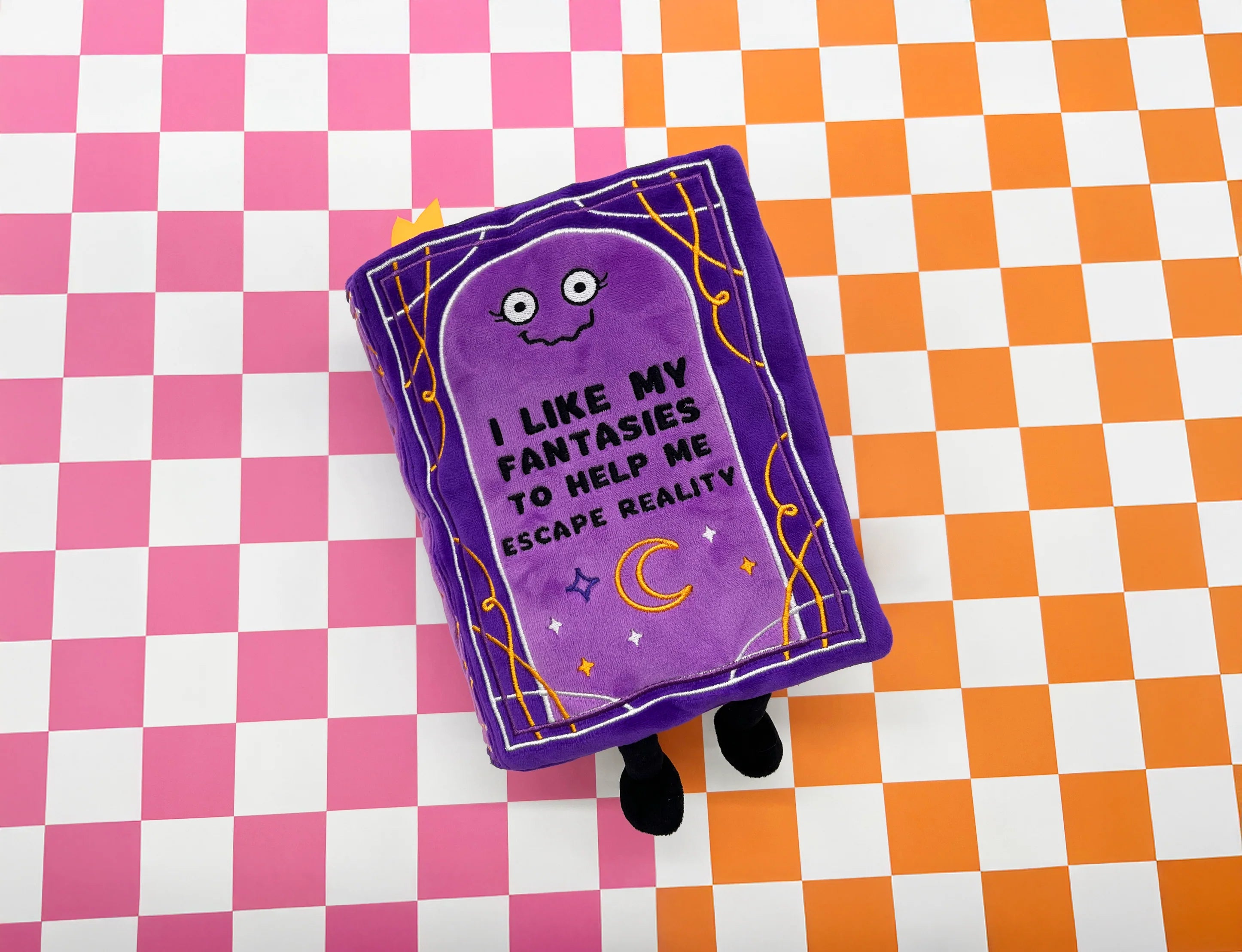 A purple plush book toy with embroidered cartoon eyes and the text 'I LIKE MY FANTASIES TO HELP ME ESCAPE REALITY' on a pink and white checkered background.