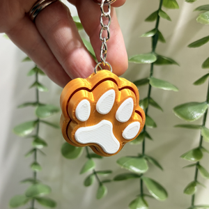 Gold keychain with a white paw print held by a hand against a green leafy background
