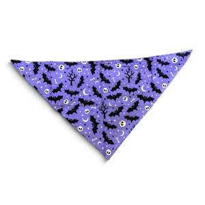 A triangular pet bandana with a purple background and a Halloween pattern featuring black bats, white skulls, and black silhouettes of trees and moons, shown on a plain white background.