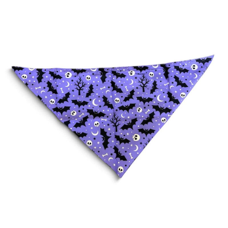 A triangular pet bandana with a purple background and a Halloween pattern featuring black bats, white skulls, and black silhouettes of trees and moons, shown on a plain white background.
