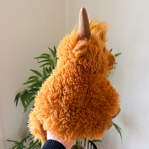 A fluffy orange Highland cow plush toy being held up by a hand, showing its textured fur and brown horn, with a green plant and white wall in the background.