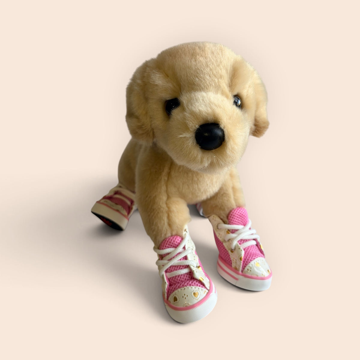 A plush golden retriever toy wearing pink and white sneakers with yellow accents, posed against a solid pale peach background.