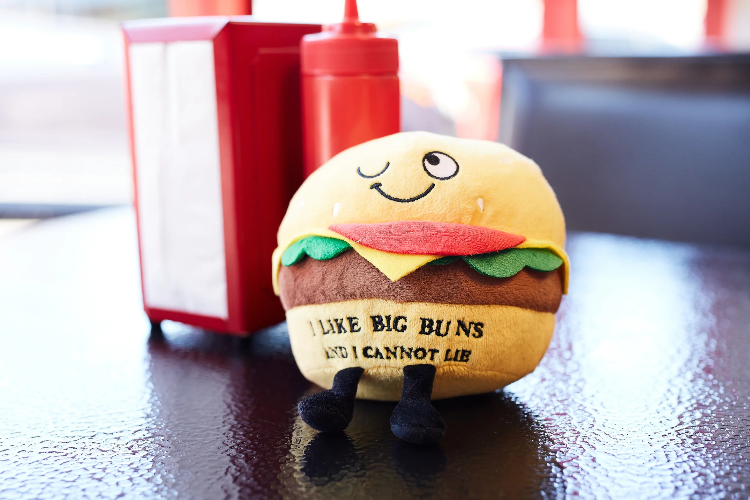 Plush burger toy with a smiley face on a table with condiment bottles in the background.
