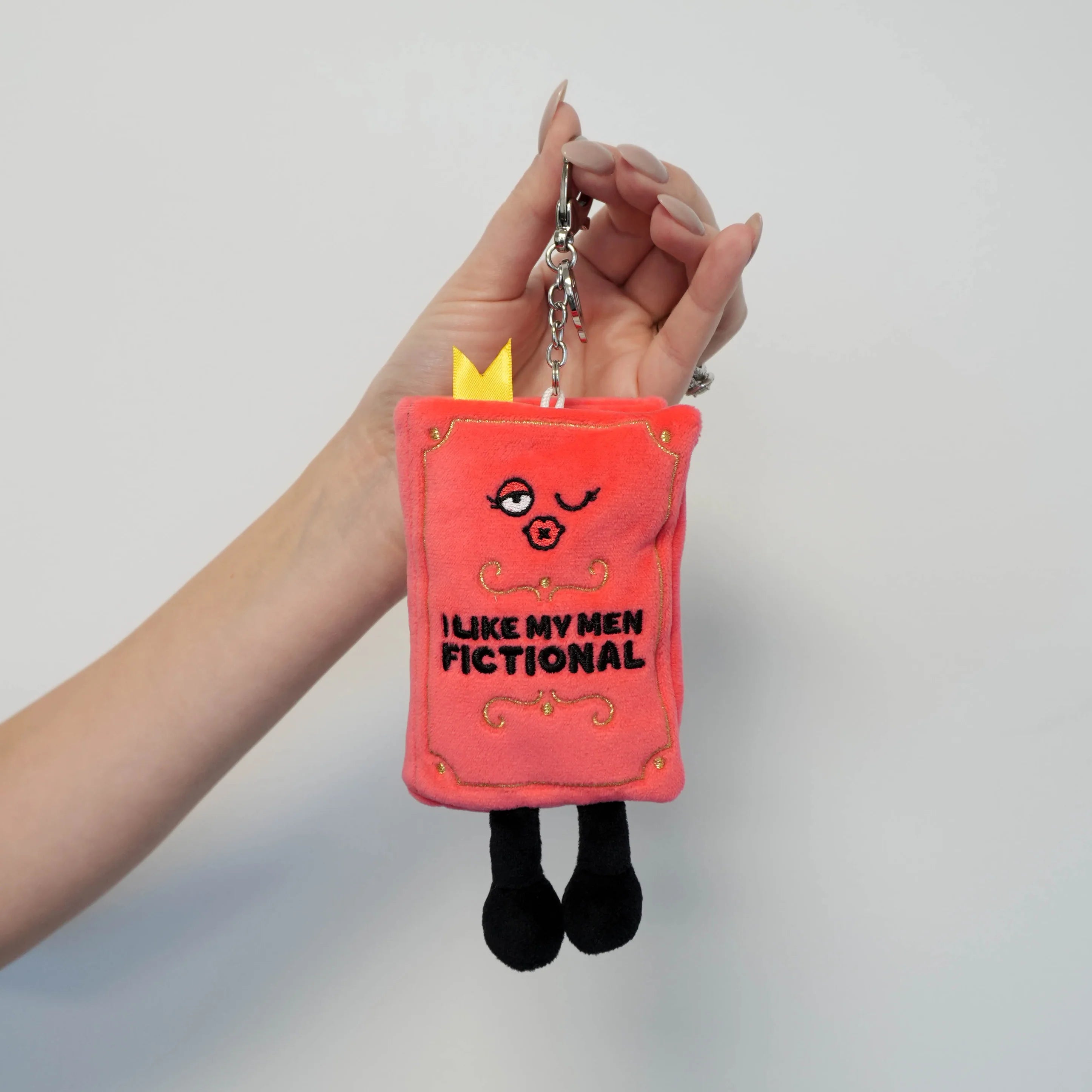 Plush keychain with cartoon face and text held by a hand against a plain background