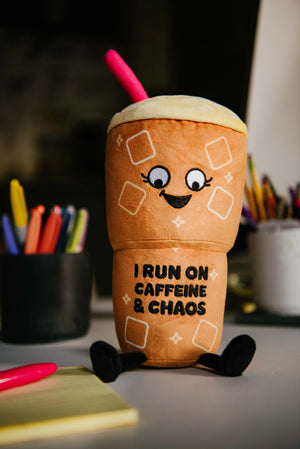 A plush toy shaped like a coffee cup with a smiling face, black legs, and a pink straw, sitting on a desk with school supplies in the background. The text on the cup reads 'I RUN ON CAFFEINE & CHAOS'.