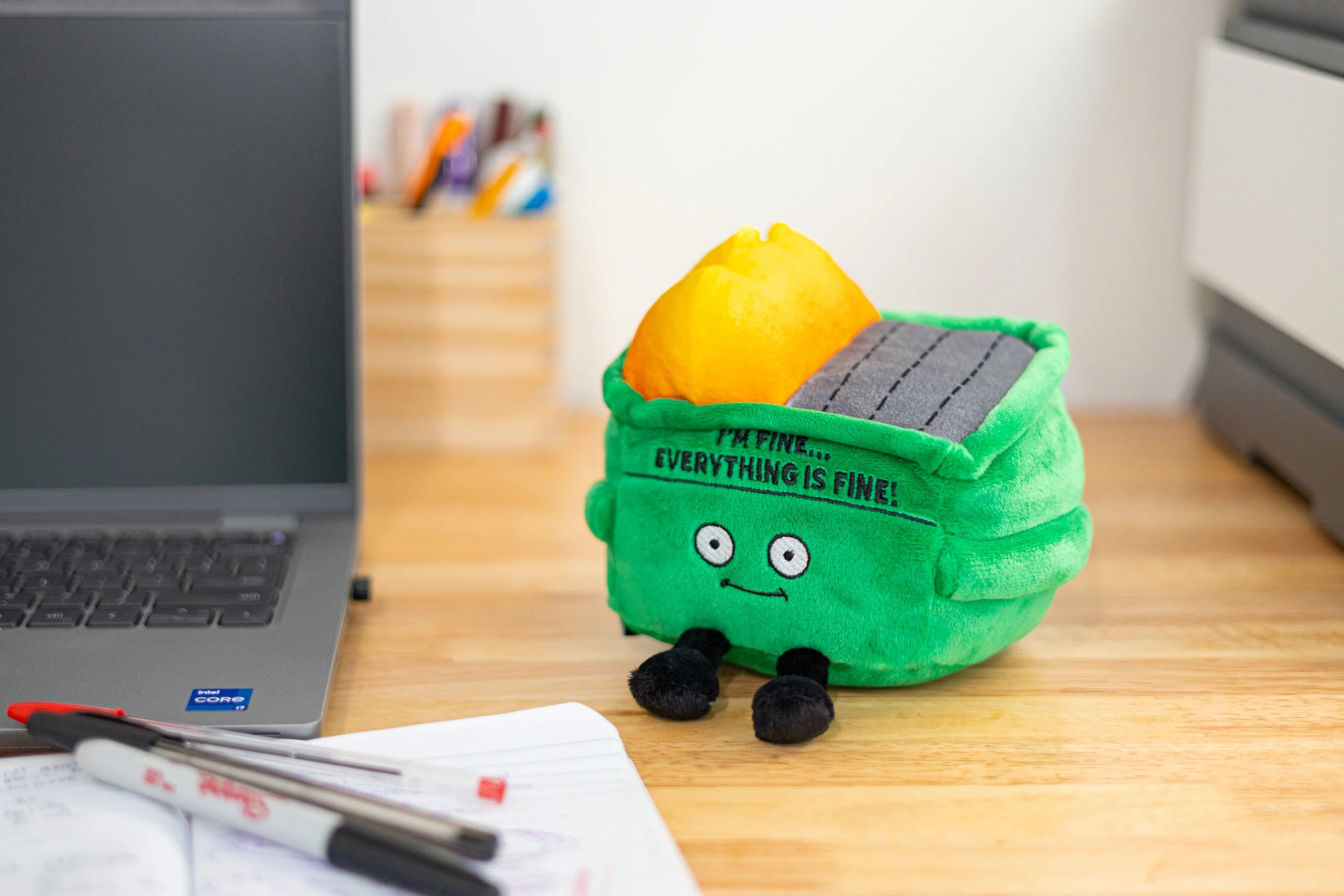 A green plush solar-panel storage bin shaped like a trash can, sitting on a wooden desk next to a laptop and notebook.