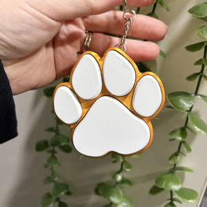 Keychain in the shape of a white paw print with gold border held by a hand against a green leafy background.