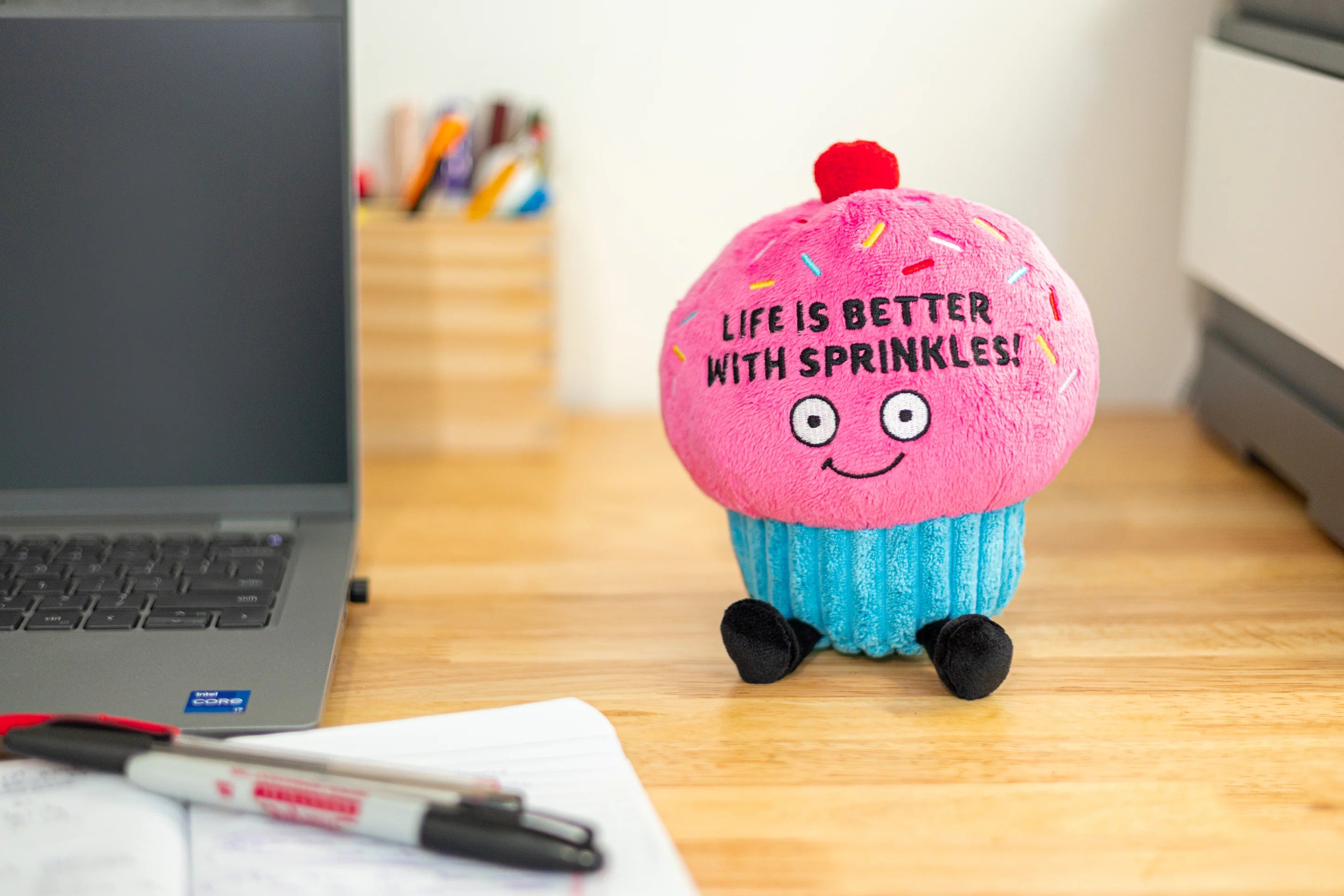 Plush cupcake toy with 'Life is better with sprinkles!' text on a desk next to a laptop.