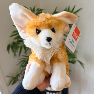A plush toy dog with large ears, orange and cream fur, and a hangtag reading Wild Animal Mini Corgi, held in a hand with a blurred indoor plant background.