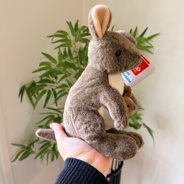 A plush gray kangaroo toy being held in a person's hand, with a small brown plush figure tucked inside its belly and a red-and-white tag attached. A green potted plant is visible in the background against a light wall.